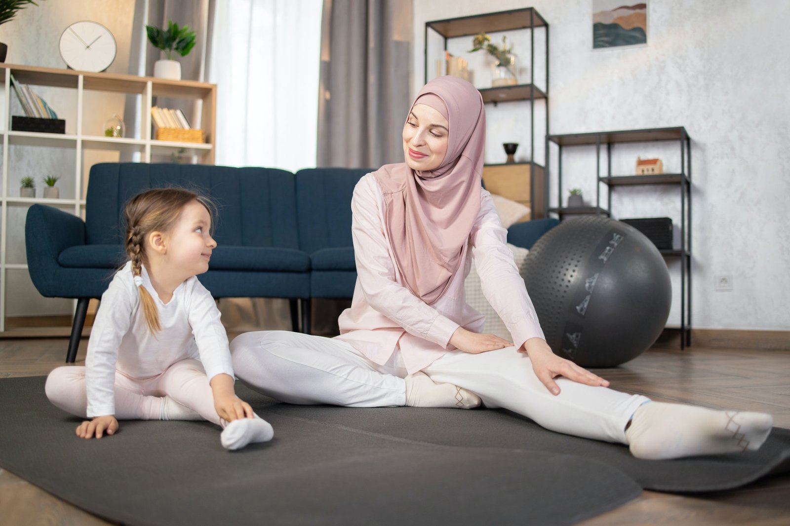 Young muslim mother in hijab showing her little daughter legs stretching exercises.