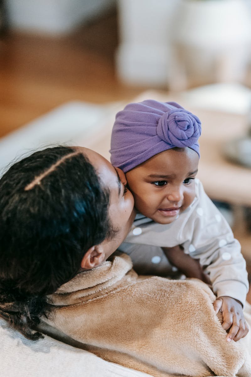 Crop ethnic mom embracing and kissing cheek of adorable little girl in headscarf at home on blurred background