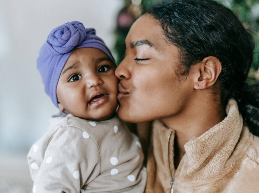 Gentle African American mum with closed eyes kissing adorable little daughter in turban at home