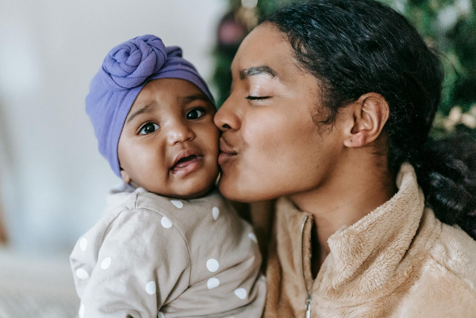 Gentle African American mum with closed eyes kissing adorable little daughter in turban at home